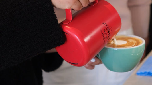 Red mug with a handle held by a person, next to a green cup with a latte.