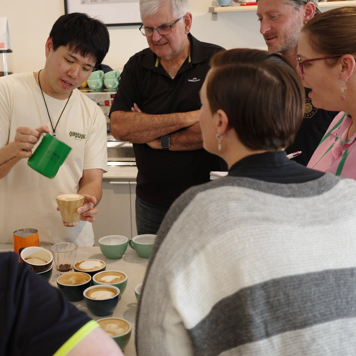 A group of people gathered around a table in a kitchen or cafe setting, with one man pouring milk from a green pitcher into a small cup. Several small bowls containing coffee drinks are arranged on the table.
